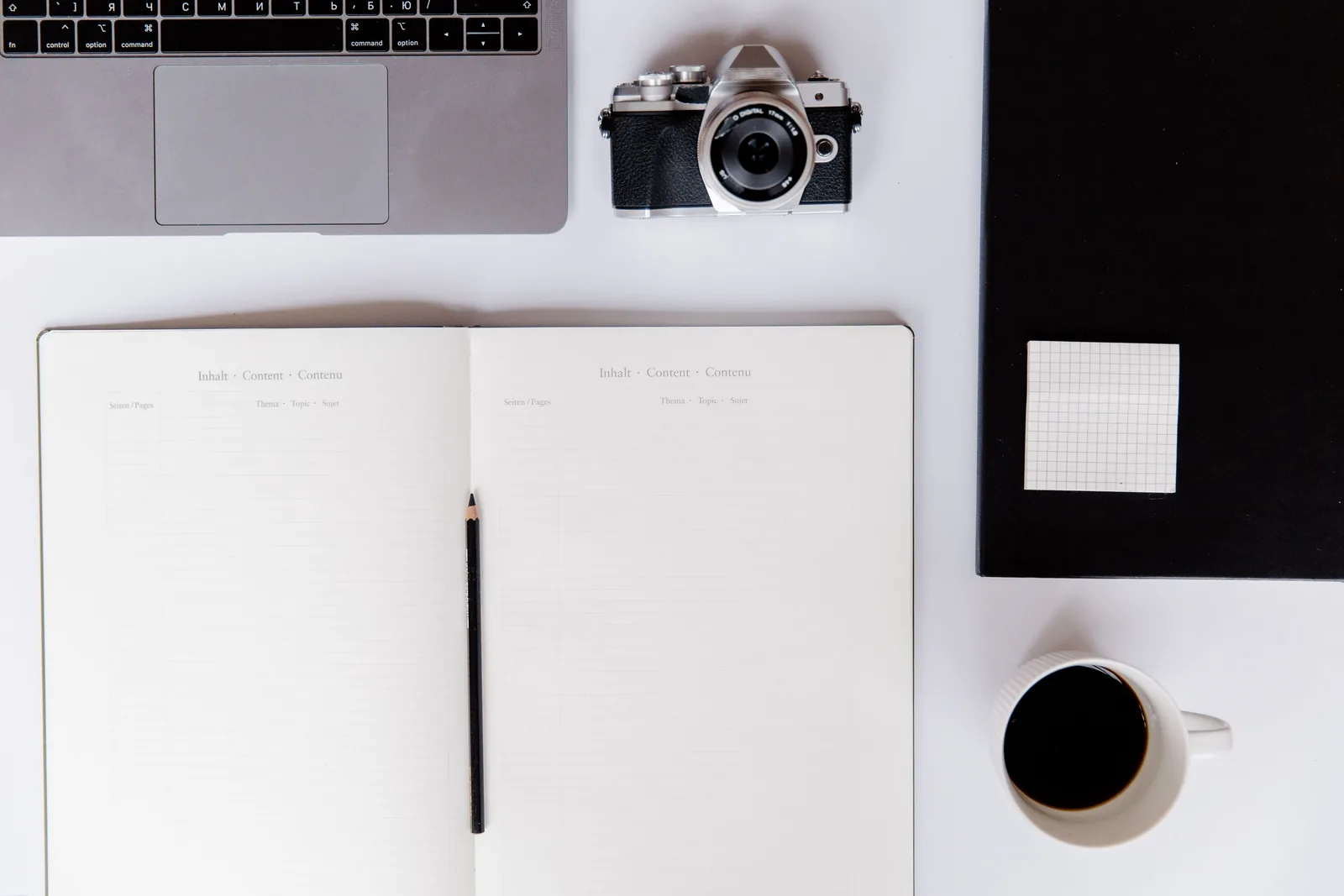 Top-down minimalist workspace with an open notebook, laptop, camera, coffee, and stationery.